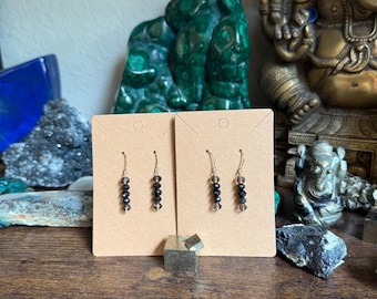 May include: Two pairs of dangle earrings with dark gray and black beads are displayed on brown earring cards. The earrings have a simple design, with the beads strung together. The background includes decorative objects and a wooden surface.