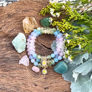 May include: A set of three beaded bracelets with a daisy charm. The bracelets are made with light blue, pink, and green beads. The charm is gold with a white daisy.