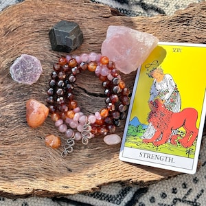 May include: A tarot card reading with the Strength card, featuring a woman with a crown of flowers and a lion. The card is surrounded by crystals, including rose quartz, carnelian, and amethyst.