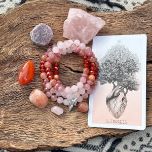 May include: A pink, red, and orange crystal bracelet with silver heart charms. The bracelet is on a wooden surface with a pink rose quartz, a red carnelian, and a tarot card with the Empress illustration. The card reads "III The Empress".