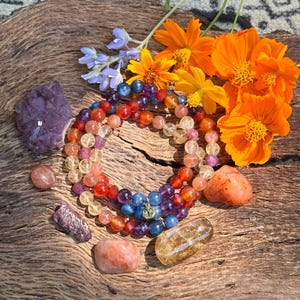 May include: A set of four gemstone bracelets with a gold charm in the center. The bracelets are made of blue, purple, red, and yellow gemstones. The bracelets are arranged on a wooden surface with orange flowers and other gemstones.