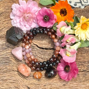 May include: A close-up of a bracelet made with various gemstones, including sunstone, amethyst, and black onyx. The bracelet is surrounded by pink and orange flowers on a wooden surface.