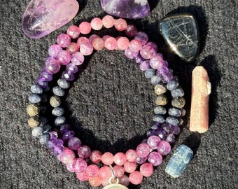 Könnte beinhalten: Eine Sammlung von Edelsteinarmbändern und Kristallen. Die Armbänder bestehen aus rosa, lila und blauen Perlen. Mehrere Amethystkristalle, ein schwarzer Stein, ein rosa Kristall und ein blauer Kristall sind um die Armbänder herum angeordnet. Ein silberner Anhänger mit Waagen-Design.