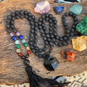 May include: A black lava stone mala necklace with a black tassel. The necklace is adorned with seven chakra crystals: amethyst, lapis lazuli, aquamarine, green aventurine, carnelian, red jasper, and black obsidian.