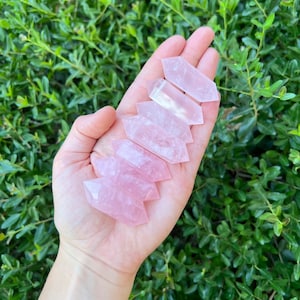 May include: Five pink rose quartz points, also known as arrowhead crystals, are arranged in a stack in a person's hand. The points are various sizes and have a natural, raw finish.