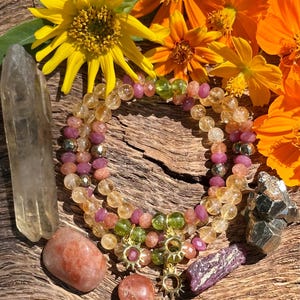 May include: A multi-strand bracelet with yellow, pink, and green beads, gold accents, and a sun charm. The bracelet is displayed on a wooden surface with sunstone crystals and a clear quartz crystal.