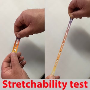 Pode incluir: Um adesivo retangular, colorido e extens&iacute;vel com a palavra "SAMPLE" impressa nele. O adesivo est&aacute; sendo esticado para mostrar sua flexibilidade.