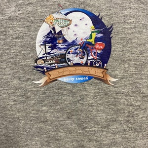 Pode incluir: Uma camiseta cinza com uma estampa gr&aacute;fica colorida de uma pessoa andando de bicicleta com um bal&atilde;o de ar quente ao fundo. O gr&aacute;fico tamb&eacute;m inclui uma lua, nuvens e um barco. O texto "party nomad" est&aacute; impresso em uma faixa.