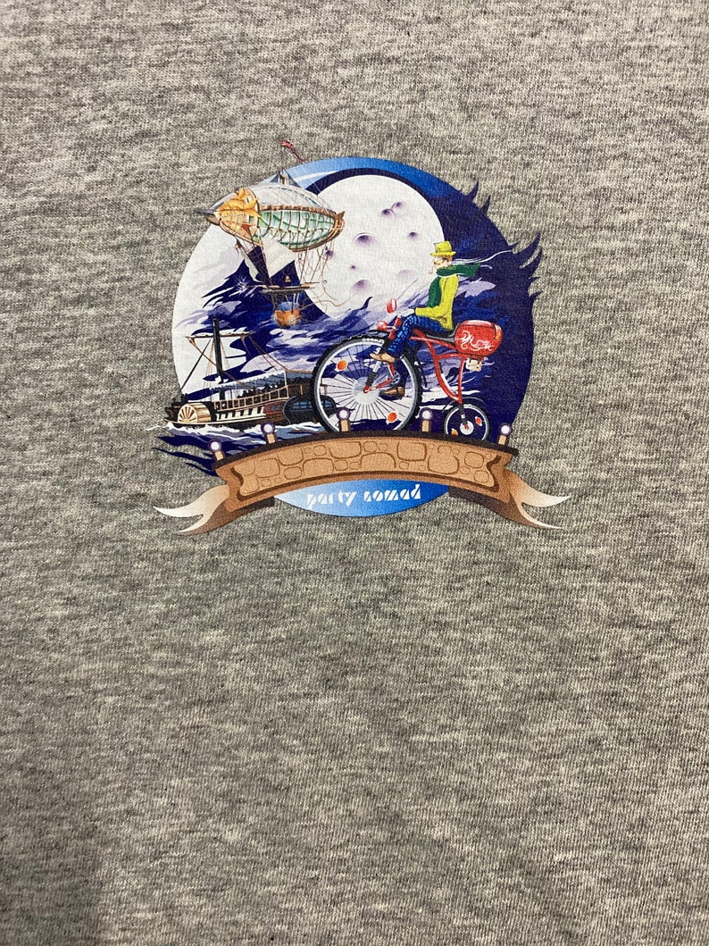 Pode incluir: Uma camiseta cinza com uma estampa gr&aacute;fica colorida de uma pessoa andando de bicicleta com um bal&atilde;o de ar quente ao fundo. O gr&aacute;fico tamb&eacute;m inclui uma lua, nuvens e um barco. O texto "party nomad" est&aacute; impresso em uma faixa.