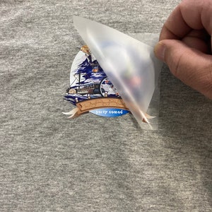 Pode incluir: Uma camiseta cinza com um transfer de ferro colorido de um navio com o texto "party sound" nela. O transfer est&aacute; sendo descascado da camiseta.