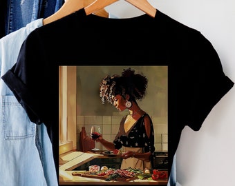 May include: A black t-shirt with a graphic print of a woman with curly hair in a kitchen setting. She is holding a glass of red wine and a plate of food. The background is a warm, inviting kitchen.