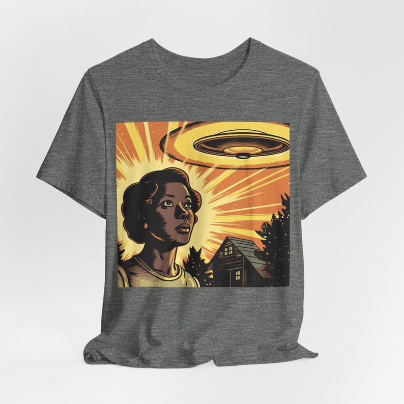 May include: A gray t-shirt with a graphic print of a woman looking up at a flying saucer. The graphic is in a retro comic book style with bright colors and bold lines. The woman is wearing a yellow shirt and has short, dark hair. The flying saucer is a classic disc shape with a bright orange glow.