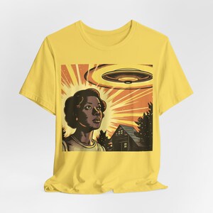 May include: A yellow t-shirt with a retro-style illustration of a person looking up at a flying saucer. The illustration is set against a background of a house and trees.