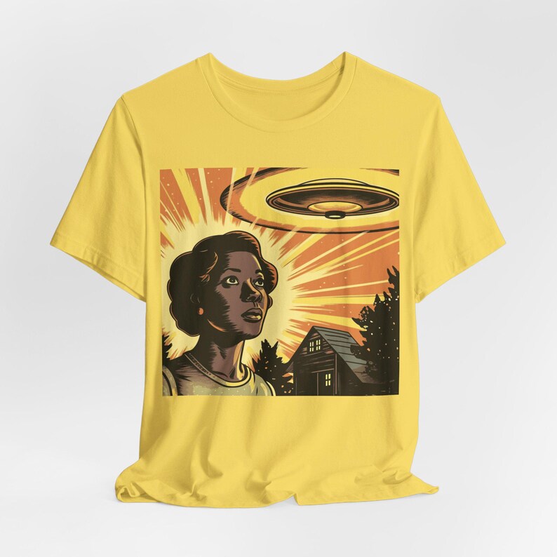 May include: A yellow t-shirt with a retro-style illustration of a person looking up at a flying saucer. The illustration is set against a background of a house and trees.