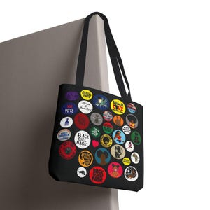 May include: Black tote bag featuring colorful circular designs with text and illustrations. The designs include phrases like "Black Girl Magic," "Black Love," and "We Vote." The bag has black handles.
