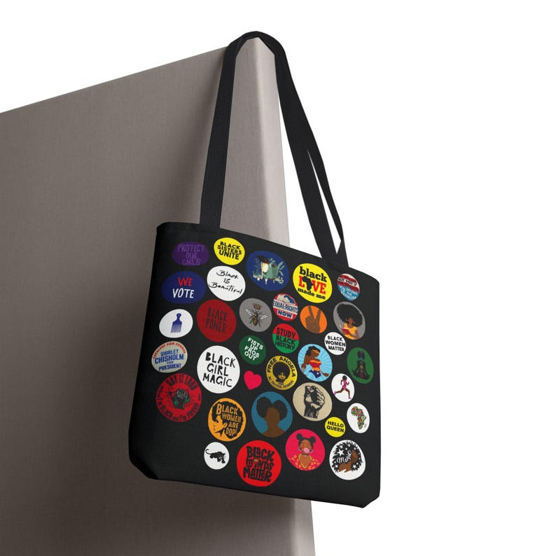 May include: Black tote bag featuring colorful circular designs with text and illustrations. The designs include phrases like "Black Girl Magic," "Black Love," and "We Vote." The bag has black handles.