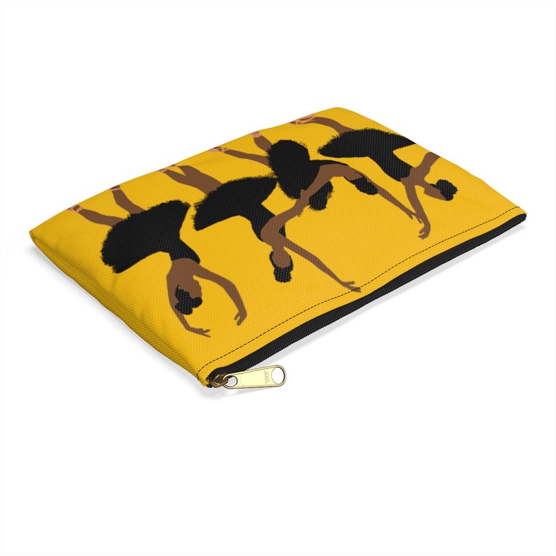 Pode incluir: Uma bolsa amarela com z&iacute;per preto, apresentando um gr&aacute;fico de tr&ecirc;s bailarinas de bal&eacute; em tutus e collants pretos. As bailarinas est&atilde;o em poses no ar contra um fundo amarelo. A bolsa tem um z&iacute;per preto e um puxador de z&iacute;per dourado.