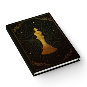 Puede incluir: Cuaderno negro con una ilustración dorada de un rey de ajedrez en la portada. El rey está representado de forma estilizada, casi abstracta, con una corona y un efecto de estrella radiante. El fondo es negro con un patrón sutil de nubes.