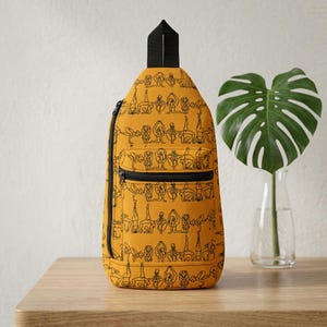 May include: Mustard yellow sling bag with a black zipper and strap. The bag features a repeating black line drawing pattern of yoga poses. The bag is on a wooden surface.