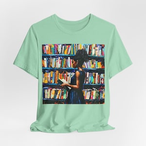 Może przedstawiać: Jasnozielony t-shirt z kolorowym nadrukiem graficznym przedstawiającym osobę stojącą przed regałem wypełnionym książkami. Osoba trzyma książkę w ręku.