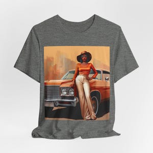 Op de afbeelding: Een grijs T-shirt met een print van een vrouw in een bruine hoed en een oranje outfit die naast een vintage oranje auto staat.