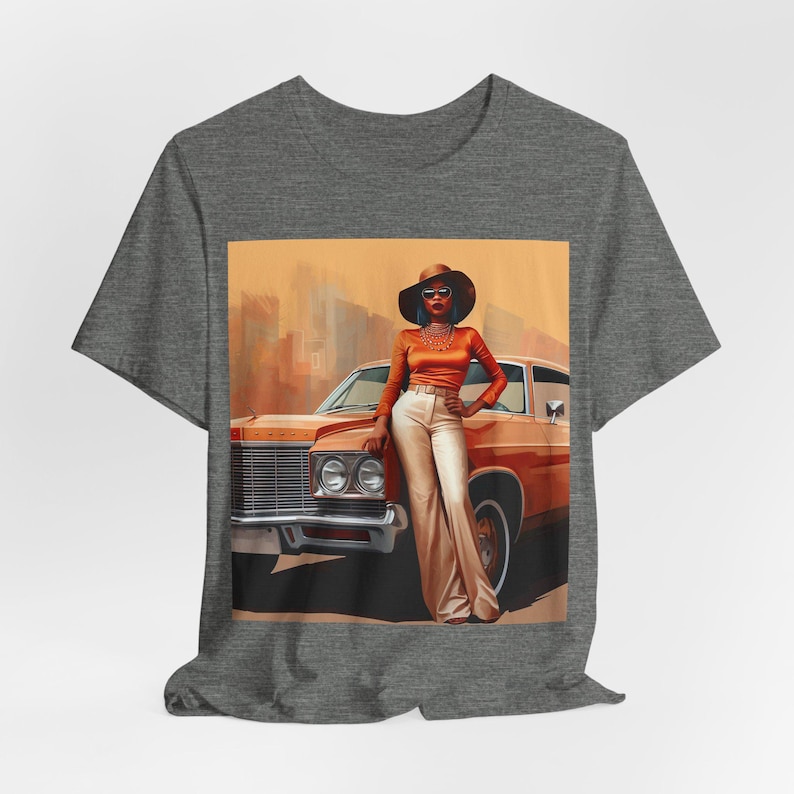 Op de afbeelding: Een grijs T-shirt met een print van een vrouw in een bruine hoed en een oranje outfit die naast een vintage oranje auto staat.