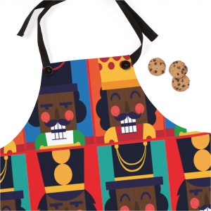 May include: A colorful kitchen apron with a repeating pattern of nutcracker faces. The nutcrackers are depicted in a cartoon style with black faces, red lips, and mustaches. The apron has a black adjustable neck strap and a black button closure.
