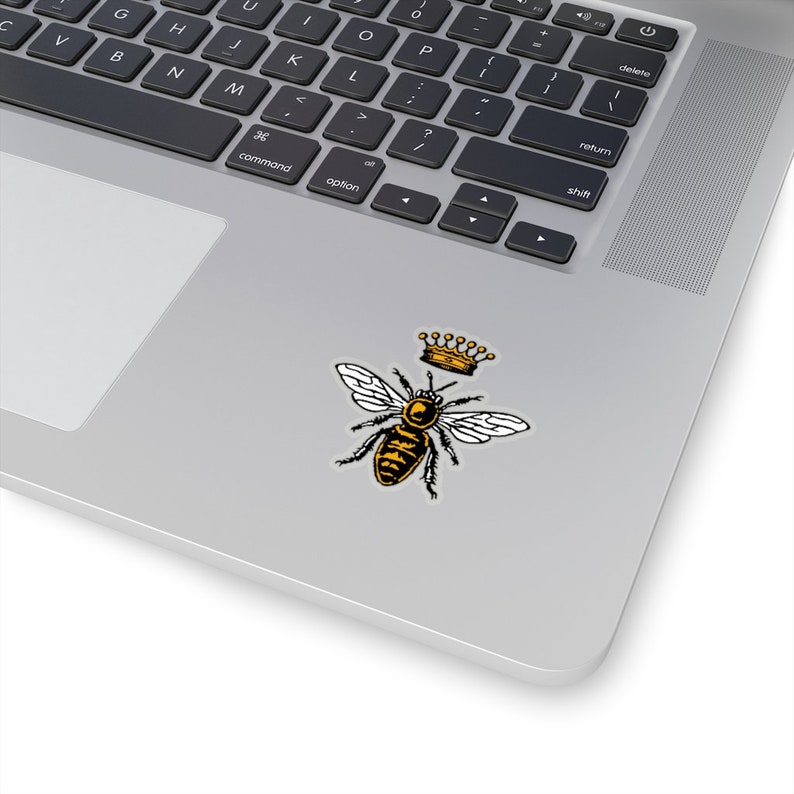 Puede incluir: Una abeja negra y amarilla con una corona dorada en la cabeza. La abeja est&aacute; en posici&oacute;n de vuelo con las alas extendidas.