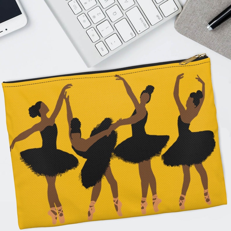 Pode incluir: Uma bolsa amarela com z&iacute;per com um gr&aacute;fico de quatro bailarinas de bal&eacute; em tutus pretos e sapatilhas de ponta rosa. As bailarinas s&atilde;o retratadas em v&aacute;rias poses graciosas em um fundo amarelo s&oacute;lido. A bolsa tem um z&iacute;per dourado.