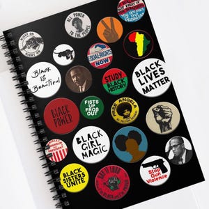 May include: A black notebook with a spiral binding features a variety of colorful buttons with black text. The buttons feature slogans such as "Black Power", "Black Lives Matter", "Black Girl Magic", "Black Sisters Unite", "Equal Rights Now", "Say It Loud", "Study Black History", "Free Angela", "Fists Up Fros Out", "Stop Gun Violence", "Right On", "All Power To The People", "Black is Beautiful", "Shirley Chisholm President".