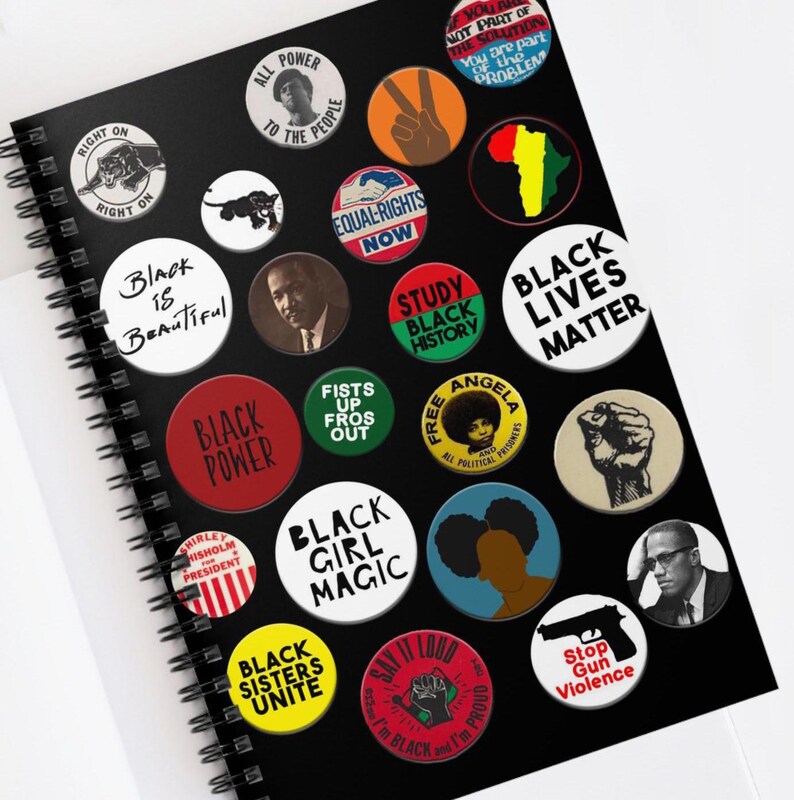 May include: A black notebook with a spiral binding features a variety of colorful buttons with black text. The buttons feature slogans such as "Black Power", "Black Lives Matter", "Black Girl Magic", "Black Sisters Unite", "Equal Rights Now", "Say It Loud", "Study Black History", "Free Angela", "Fists Up Fros Out", "Stop Gun Violence", "Right On", "All Power To The People", "Black is Beautiful", "Shirley Chisholm President".