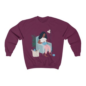 May include: A maroon sweatshirt with a graphic of a person sitting in a chair knitting. The person has dark skin and is wearing a blue and white polka dot shirt and pink pants. There is a lamp and a plant in the background.