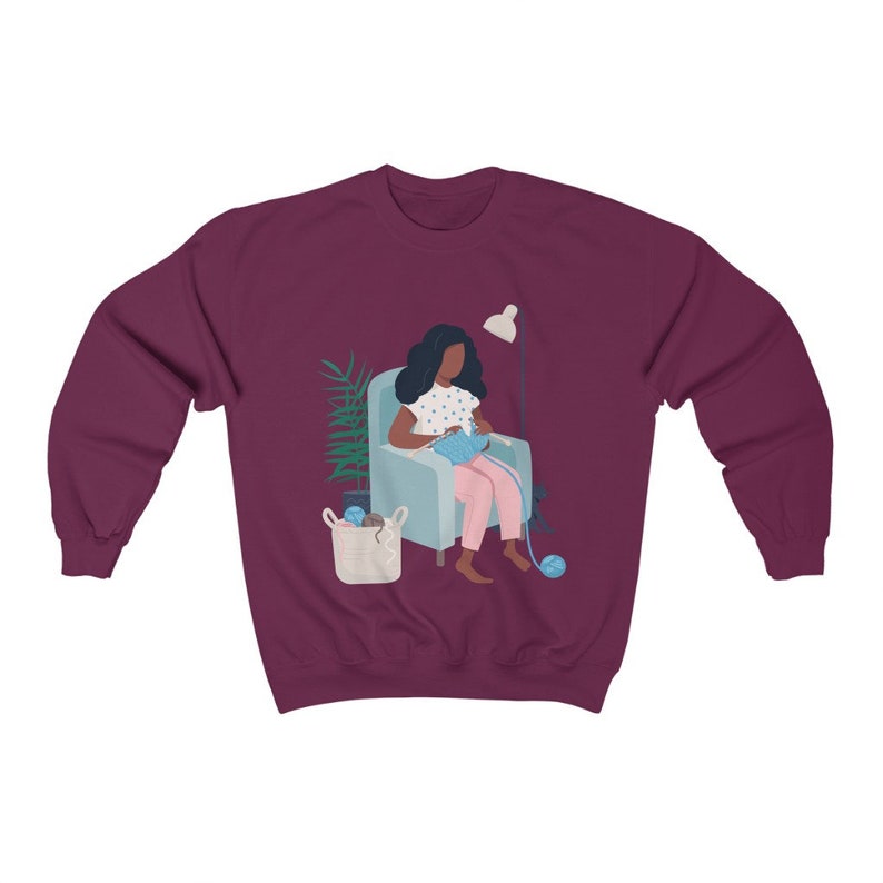 May include: A maroon sweatshirt with a graphic of a person sitting in a chair knitting. The person has dark skin and is wearing a blue and white polka dot shirt and pink pants. There is a lamp and a plant in the background.