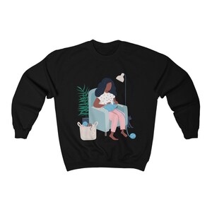 May include: A black sweatshirt with a graphic of a person sitting in a chair knitting with a cat beside them. The person is wearing a pink dress and has dark hair. There is a plant and a basket of yarn in the background.