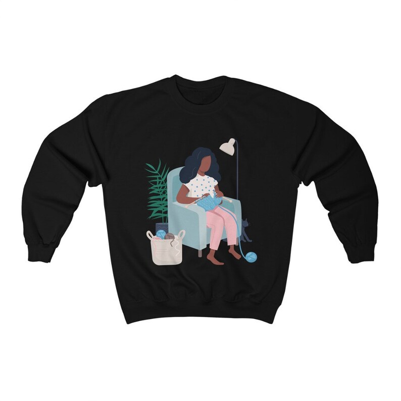 May include: A black sweatshirt with a graphic of a person sitting in a chair knitting with a cat beside them. The person is wearing a pink dress and has dark hair. There is a plant and a basket of yarn in the background.