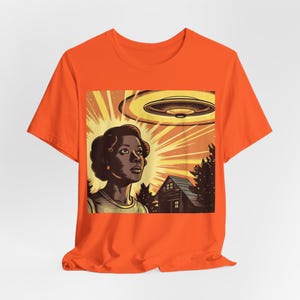 May include: Orange t-shirt with a graphic print of a woman looking up at a flying saucer. The graphic also includes a house and trees.