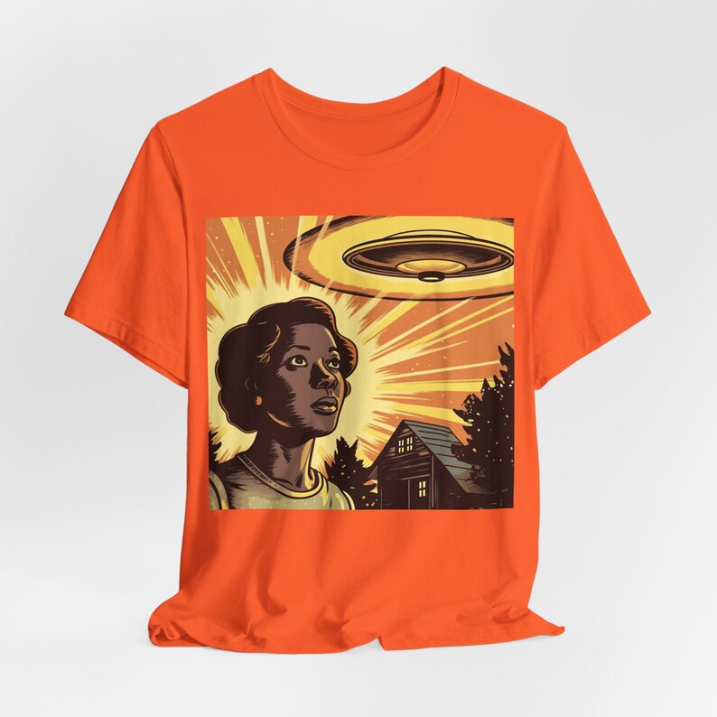 May include: Orange t-shirt with a graphic print of a woman looking up at a flying saucer. The graphic also includes a house and trees.