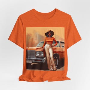 Op de afbeelding: Oranje T-shirt met een grafische print van een vrouw in een bruine hoed en zonnebril die naast een vintage auto staat.
