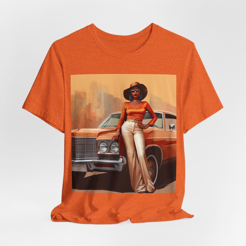 Op de afbeelding: Oranje T-shirt met een grafische print van een vrouw in een bruine hoed en zonnebril die naast een vintage auto staat.