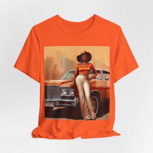Op de afbeelding: Oranje T-shirt met een grafische print van een vrouw in een bruine auto. De vrouw draagt een bruine hoed, een zonnebril en een bruin en oranje outfit. De auto is een vintage model met een bruine buitenkant.