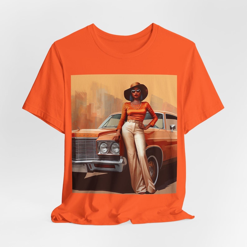 Op de afbeelding: Oranje T-shirt met een grafische print van een vrouw in een bruine auto. De vrouw draagt een bruine hoed, een zonnebril en een bruin en oranje outfit. De auto is een vintage model met een bruine buitenkant.