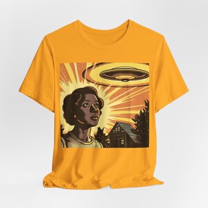 May include: A yellow t-shirt with a graphic print of a woman looking up at a flying saucer. The graphic also includes a house and trees in the background.