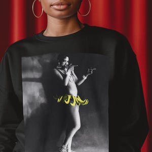 May include: Black sweatshirt featuring a graphic print of a woman wearing a yellow banana skirt and a pearl necklace. The woman is pointing to the side and smiling.