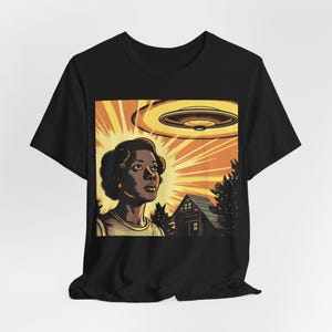 May include: A black t-shirt with a graphic print of a woman looking up at a flying saucer. The woman is standing in front of a house with trees in the background. The graphic is in a retro style with bright colors.