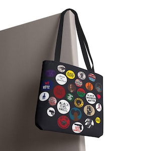 May include: Black tote bag featuring colorful circular designs with text like "Black Girl Magic", "Black Women Matter", and "We Vote". The bag has black handles and is hanging against a neutral background.
