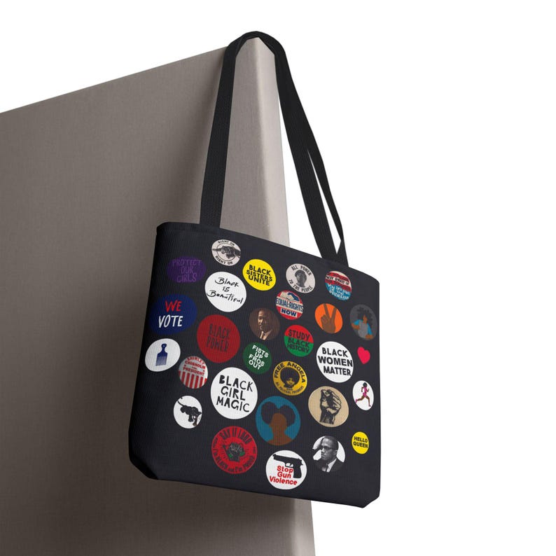 May include: Black tote bag featuring colorful circular designs with text like "Black Girl Magic", "Black Women Matter", and "We Vote". The bag has black handles and is hanging against a neutral background.
