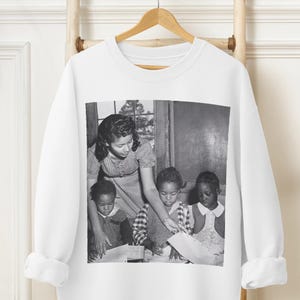 May include: Black and white photo of a woman teaching three children in a classroom. The woman is standing and the children are sitting at desks. The woman is wearing a dress and the children are wearing dresses. The photo is printed on a white sweatshirt.