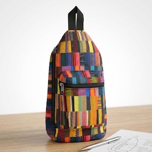 May include: A vertical sling backpack featuring a vibrant, multi-colored patchwork pattern of rectangular stripes in shades of yellow, orange, red, blue, and green. The bag includes a black top handle, a front zippered pocket, and a side zipper closure.