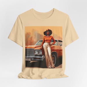 Op de afbeelding: Een beige T-shirt met een grafische print van een vrouw met een bruine hoed en zonnebril, die naast een oranje vintage auto staat.