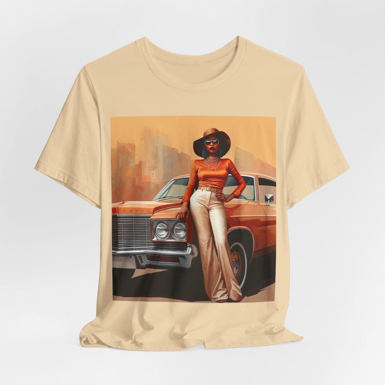 Op de afbeelding: Een beige T-shirt met een grafische print van een vrouw met een bruine hoed en zonnebril, die naast een oranje vintage auto staat.